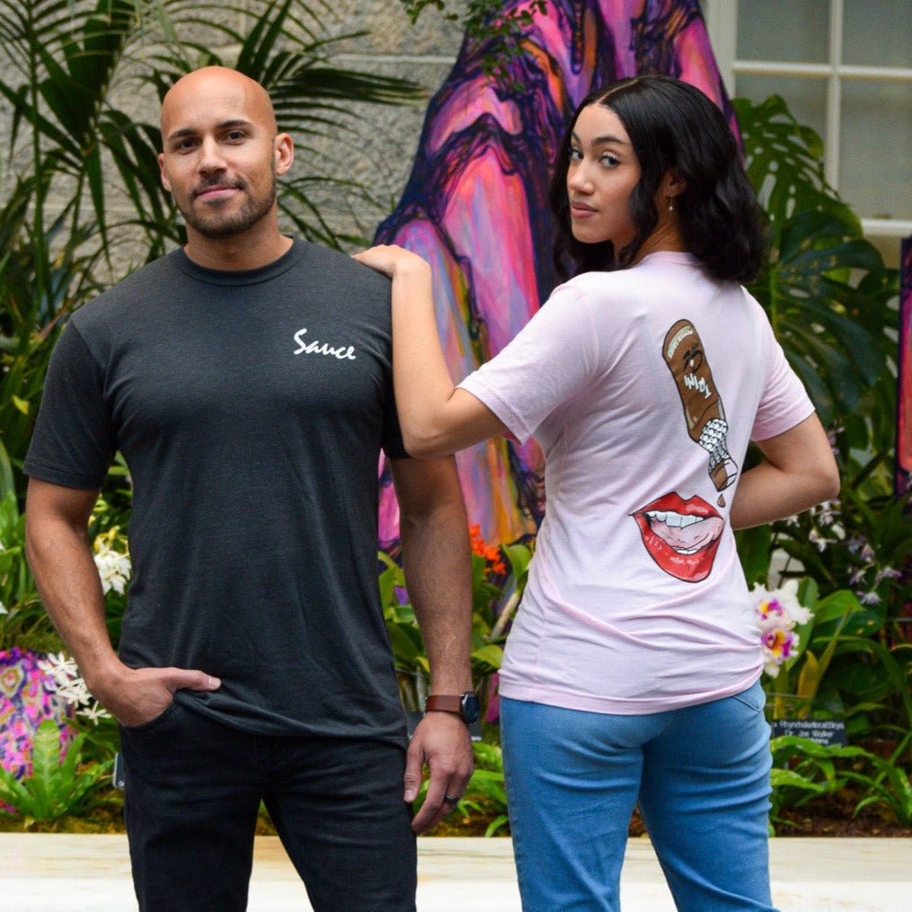Two models with the Trini Pepper sauce shirt. The male, in grey, showing the front. The female is looking over her shoulder showing the back of the shirt, The front of the shirt has sauce in cursive on the left chest of the shirt. The back of the shirt has a drawing of the Trini Sauce bottle dripping sauce onto a tongue that is sticking out from the lips and a mouth. Plants in the background.