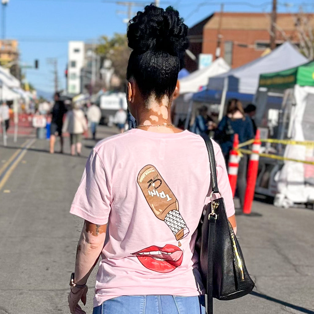 Model facing away wearing the pink Trini Sauce Shirt. The back of the shirt has a drawing of the Trini Sauce bottle dripping sauce onto a tongue that is sticking out from the lips and a mouth. They are standing in the middle of a road that has vending tents lining both sides.